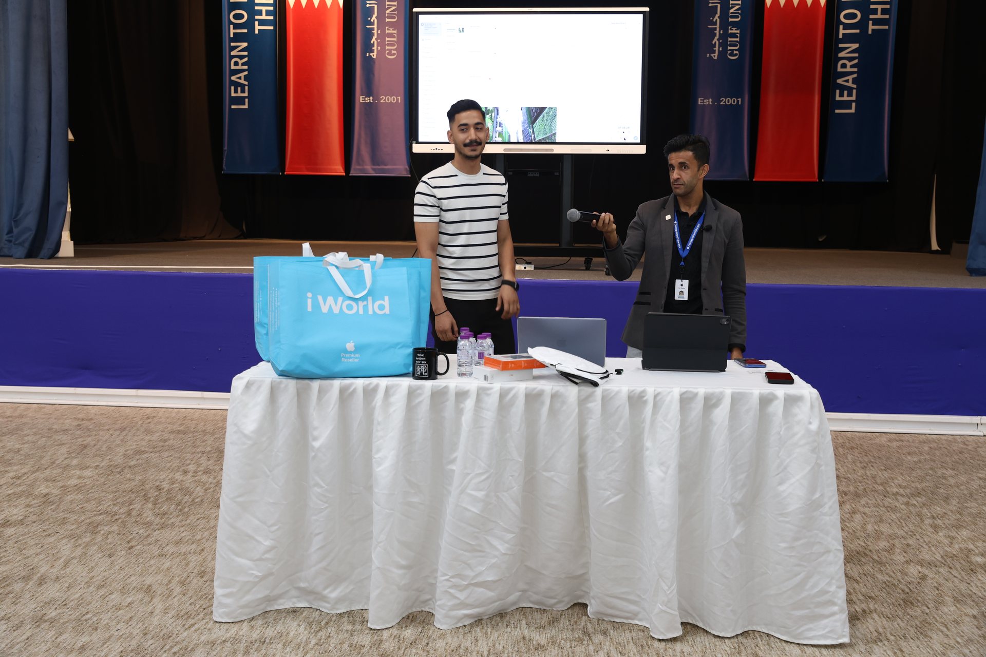 Presenters at the Gulf University and iWorld MacBook workshop standing beside a table with laptops and iWorld gift bags, in front of the Gulf University 25th anniversary banners