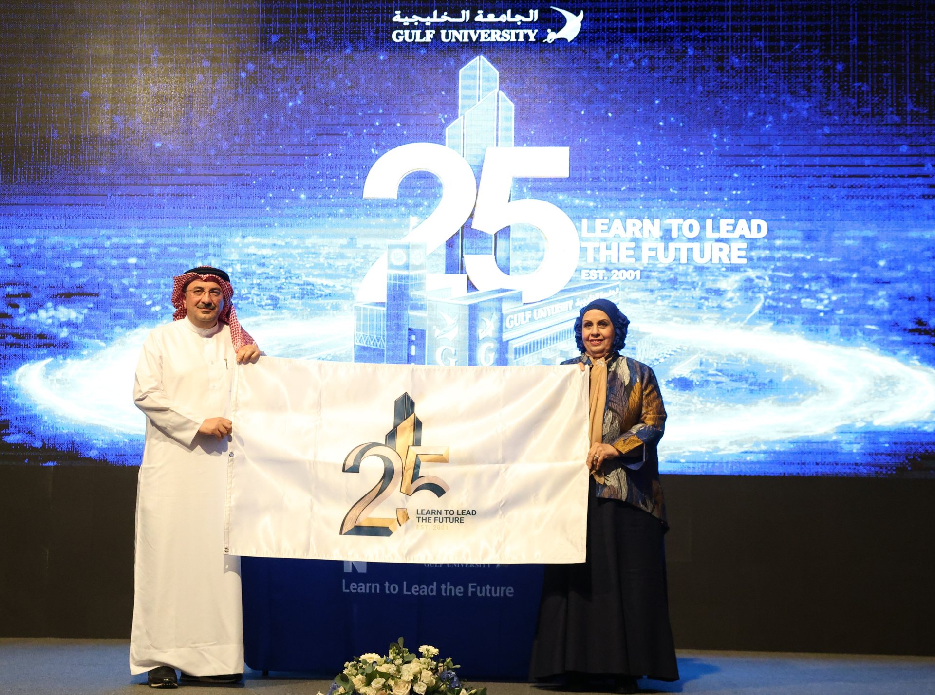 Gulf University leadership unveiling the Silver Jubilee flag bearing the 25th anniversary emblem and the slogan “Learn to Lead the Future”, on stage in front of a large screen showing the anniversary logo